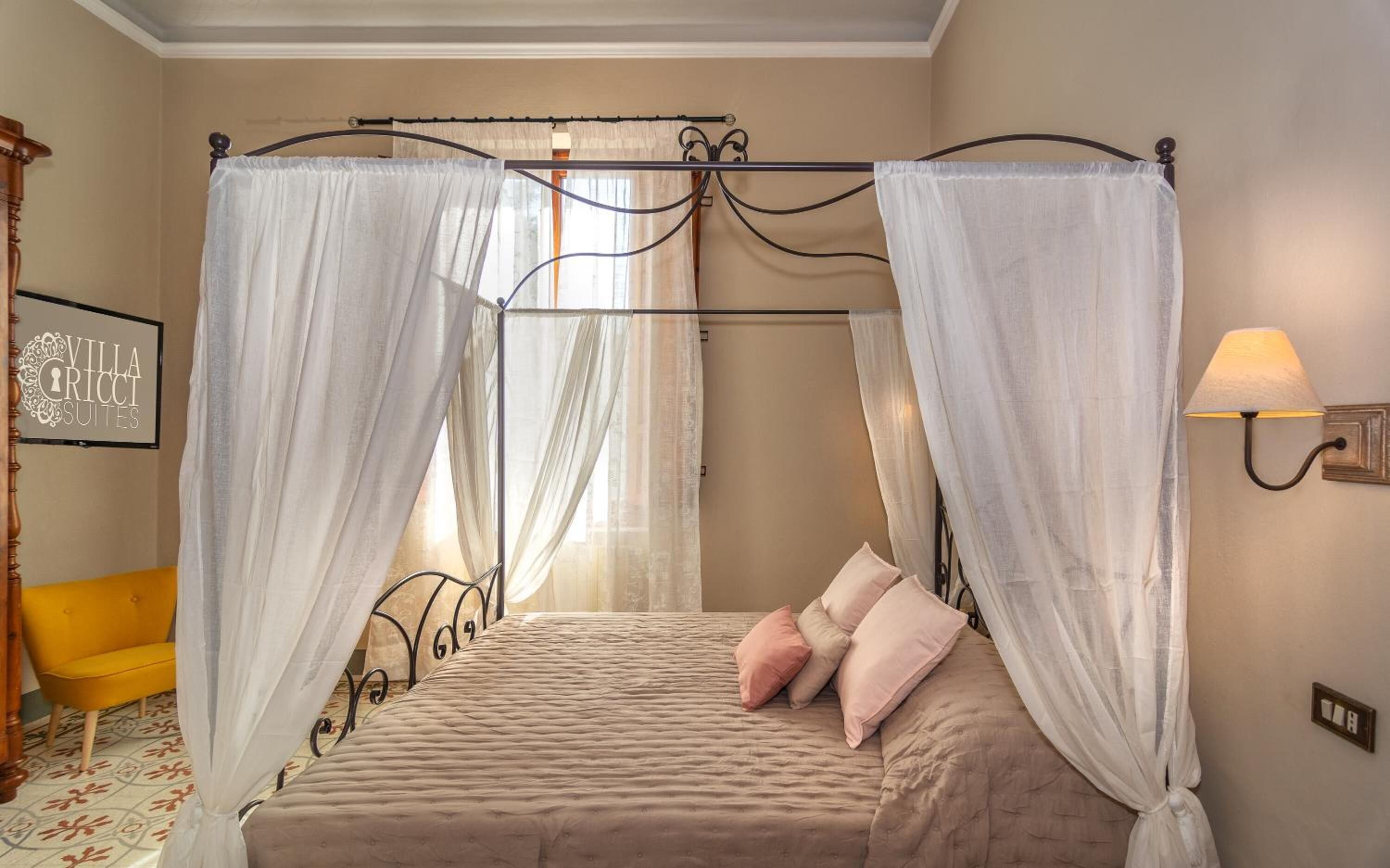 A four-poster canopy bed with a beige bedspread and pink pillows, a yellow armchair, and a wall-mounted lamp.