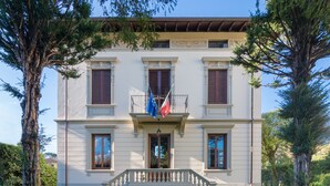 A two-story building with a symmetrical facade, a central entrance with a balustrade, and a flag on the balcony. - Villa Ricci Suites (Lucca)