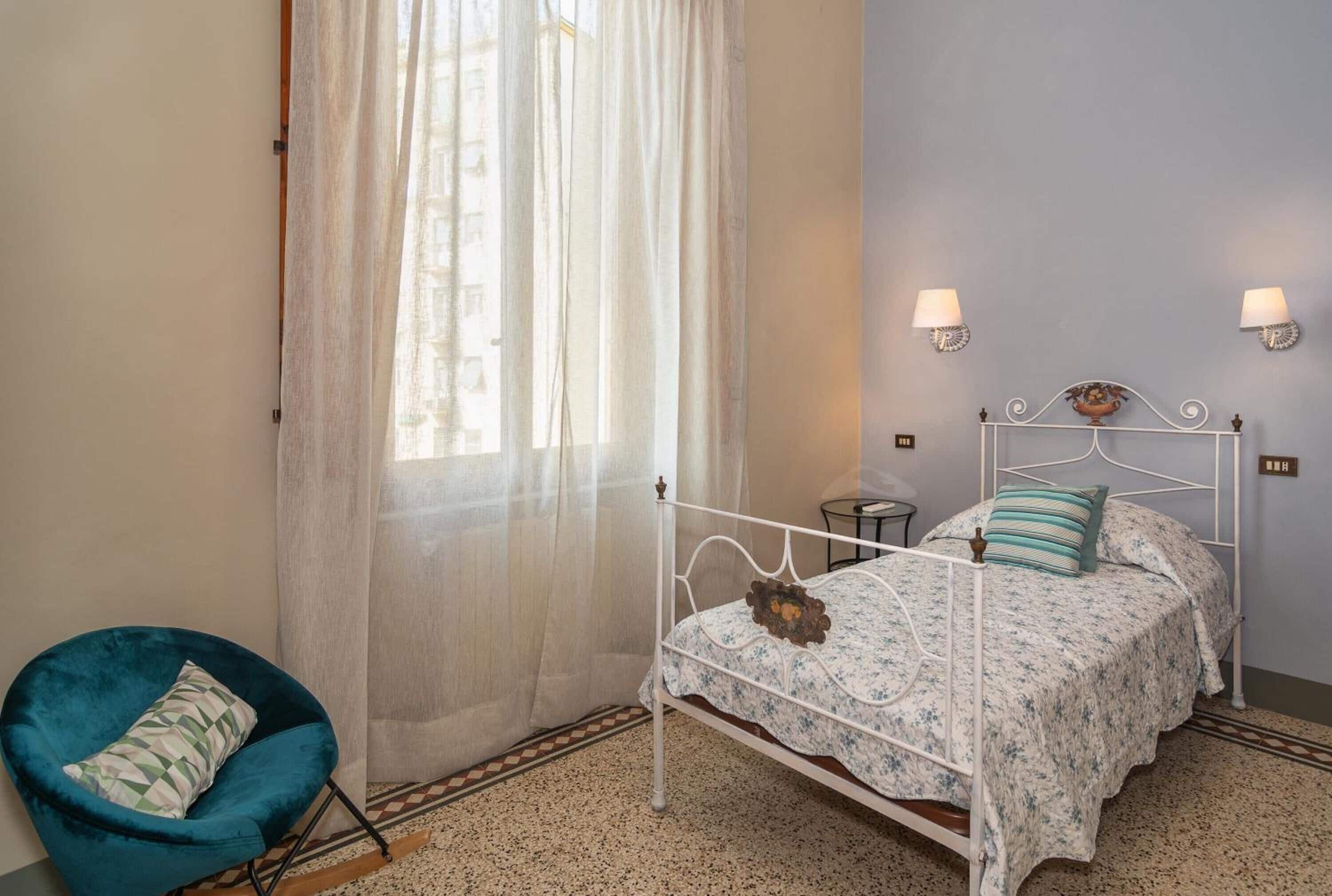 Een slaapkamer met een bed, een stoel en een raam met dunne gordijnen.