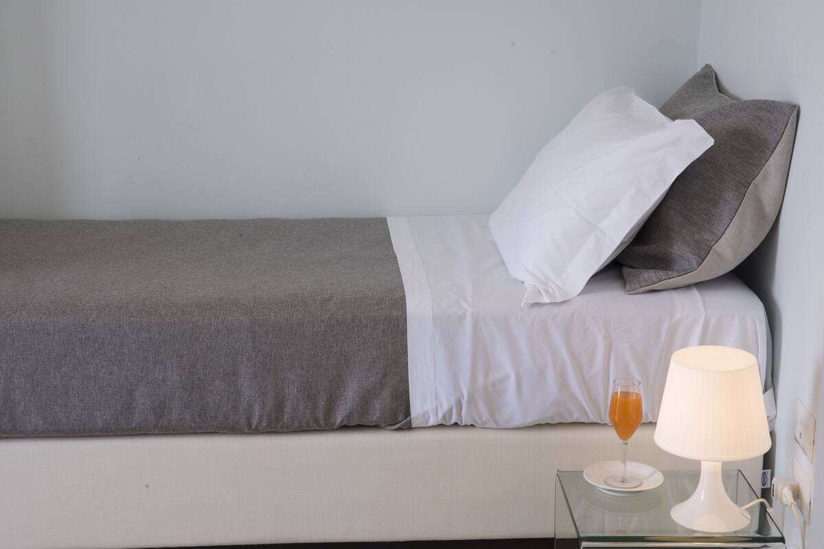 A neatly made bed with a grey blanket and white sheets, a glass of orange drink on a coaster, and a white table lamp beside it.