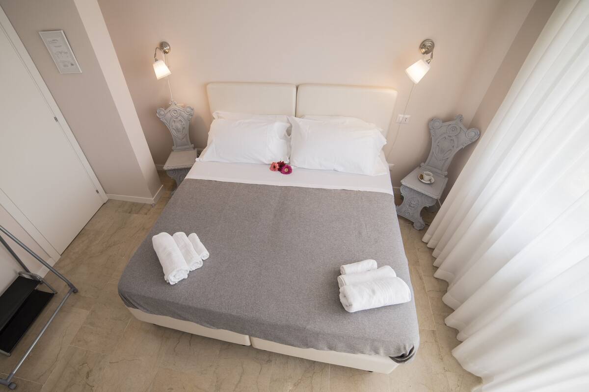A neatly made bed with white linens and towels, a grey bedspread, and two bedside tables with lamps.