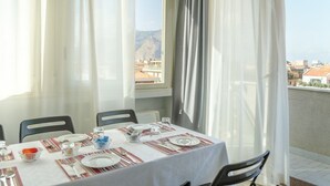 Una mesa de comedor dispuesta con platos, cubiertos y vasos, con vistas a edificios y montañas a través de cortinas translúcidas.