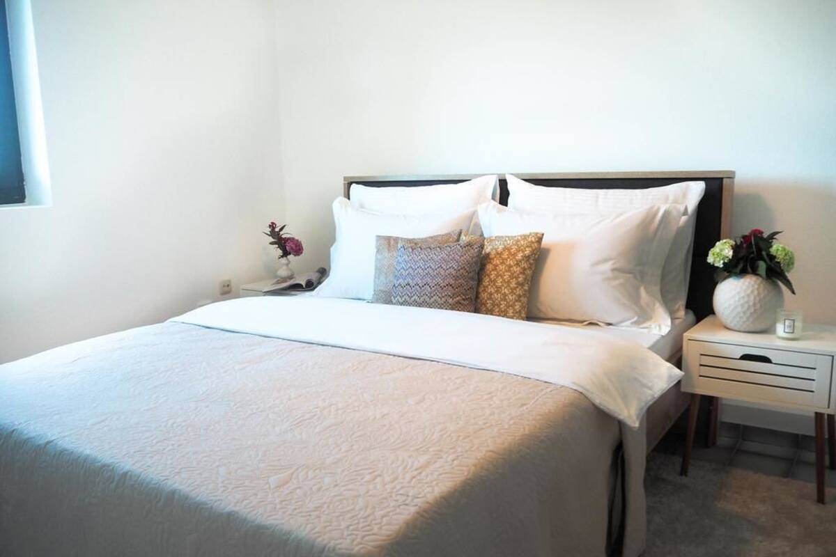 A neatly made bed with a white duvet, a wooden headboard, and a bedside table with a vase of flowers.