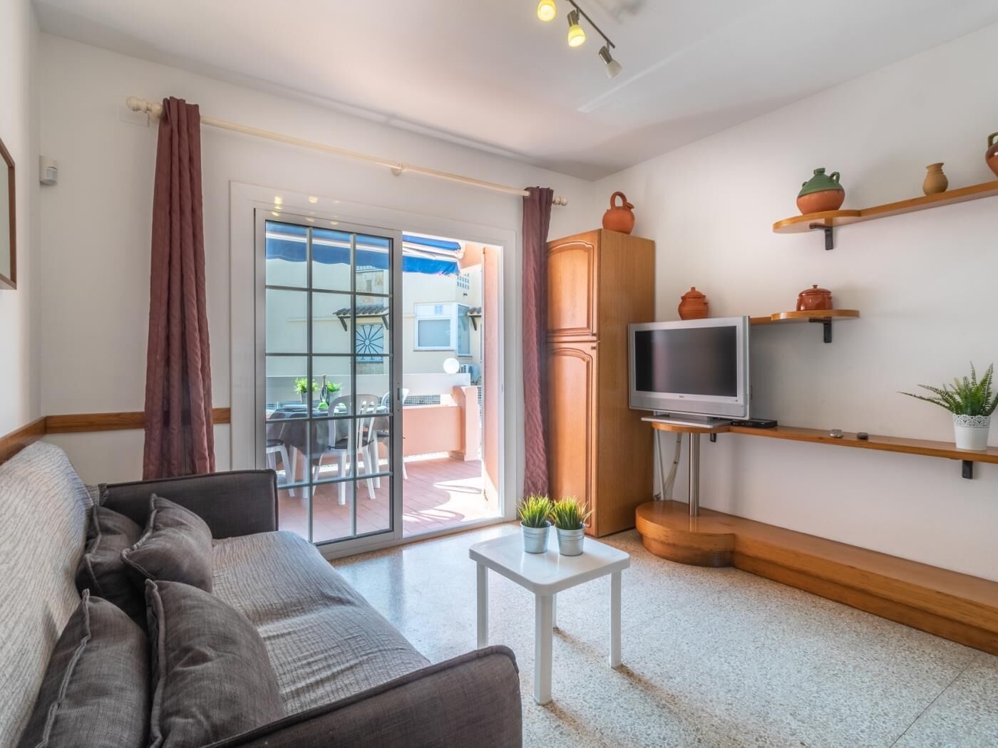 Un salon moderne avec un canapé, un téléviseur posé sur un meuble, une petite table avec des plantes et une porte-fenêtre coulissante donnant sur un espace extérieur.