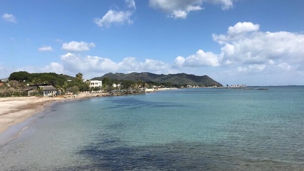 A coastal beach with clear water, a sandy shore, and a few buildings in the background. - Single villa from Titti and Carlo a stone's throw from the sea (Perd'e Sali)