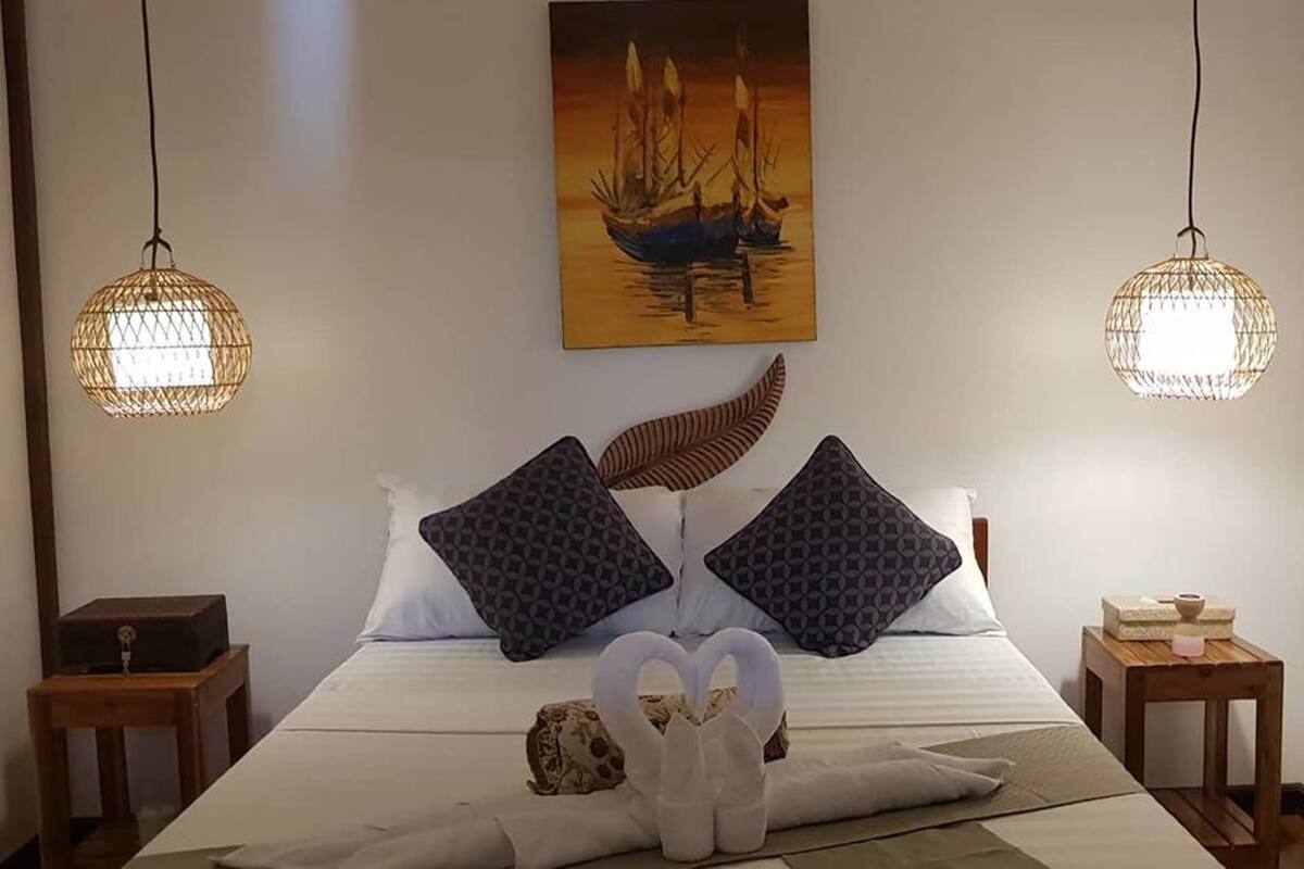 A neatly made bed with patterned pillows, two bedside tables, a painting of a boat on the wall, and two hanging lamps.