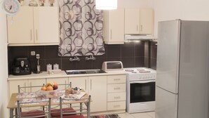A compact kitchen with a refrigerator, stove, microwave, and a coffee maker. There is a dining table with chairs and a fruit bowl. - Sofia's Residence by Estia (Heraklion)