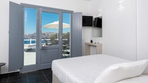 A bedroom with a bed, a TV mounted on the wall, and a sliding glass door leading to an outdoor area with a chair and umbrella. - Pancratium villas & suites (Santorini)