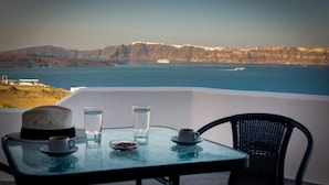 A table with a hat, two glasses, and a cup on a balcony overlooking a coastal view with mountains and a ship in the distance. - Pancratium villas & suites (Santorini)