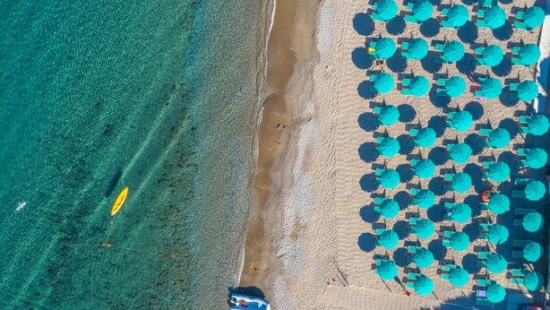 Luchtbeeld van een strand met helder turquoise water, een zandstrand en vele blauwe parasols in rijen.
