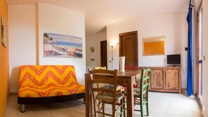 Een gezellige woonkamer met een bank, een houten eettafel, stoelen, en een keukenhoek met kastjes en een televisie.