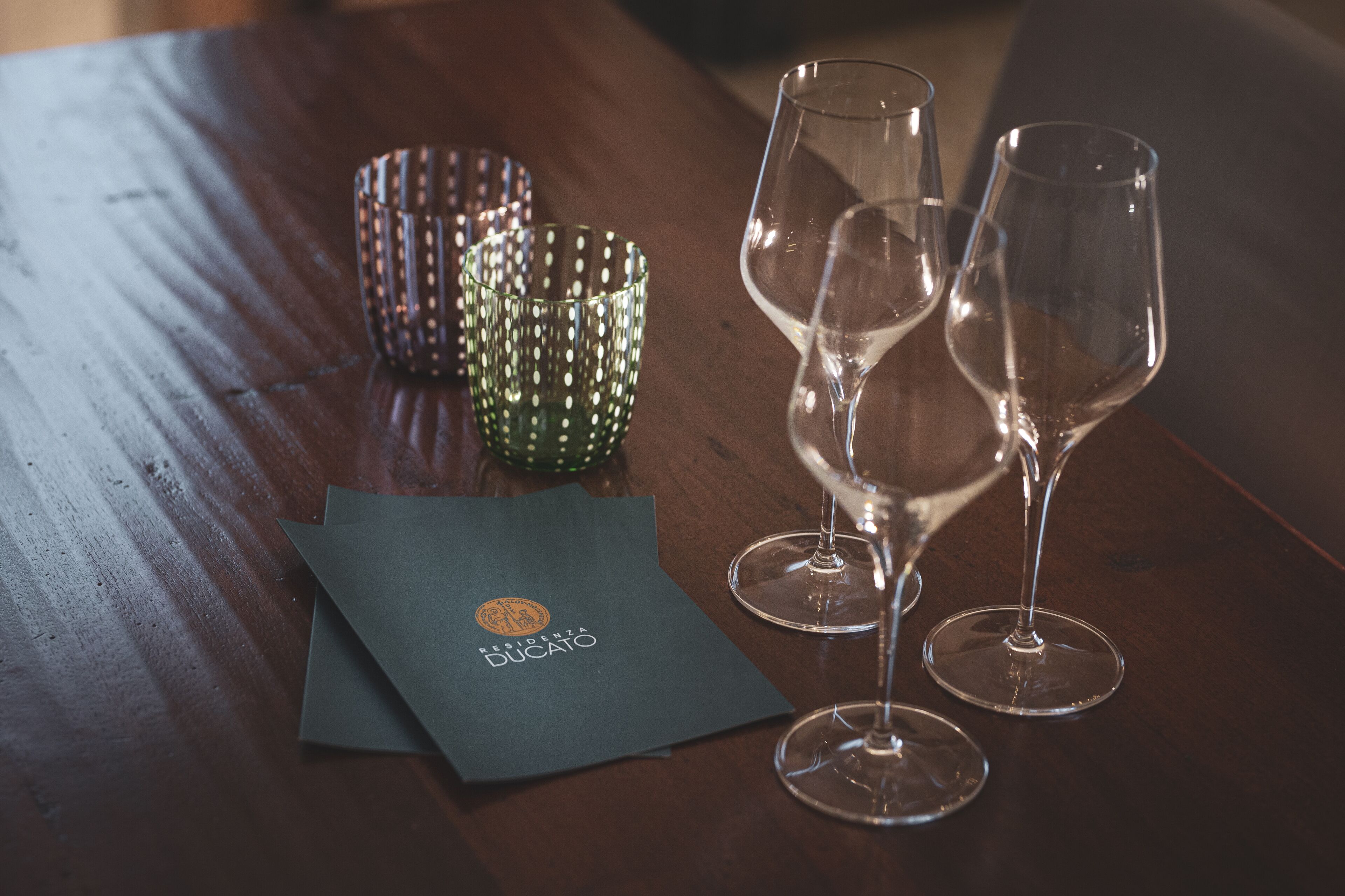 A wooden table with two empty wine glasses and a green patterned candle holder. A dark-colored envelope with a logo is also visible.