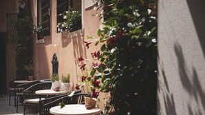 Un café en plein air avec des tables et des chaises rondes, des plantes en pot et un bâtiment dont les fenêtres sont protégées par des barreaux.