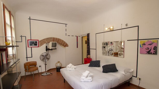 A bedroom with a bed, a chair, a fan, a TV, and a mirror on the wall. - Porcellana Art (Florence)