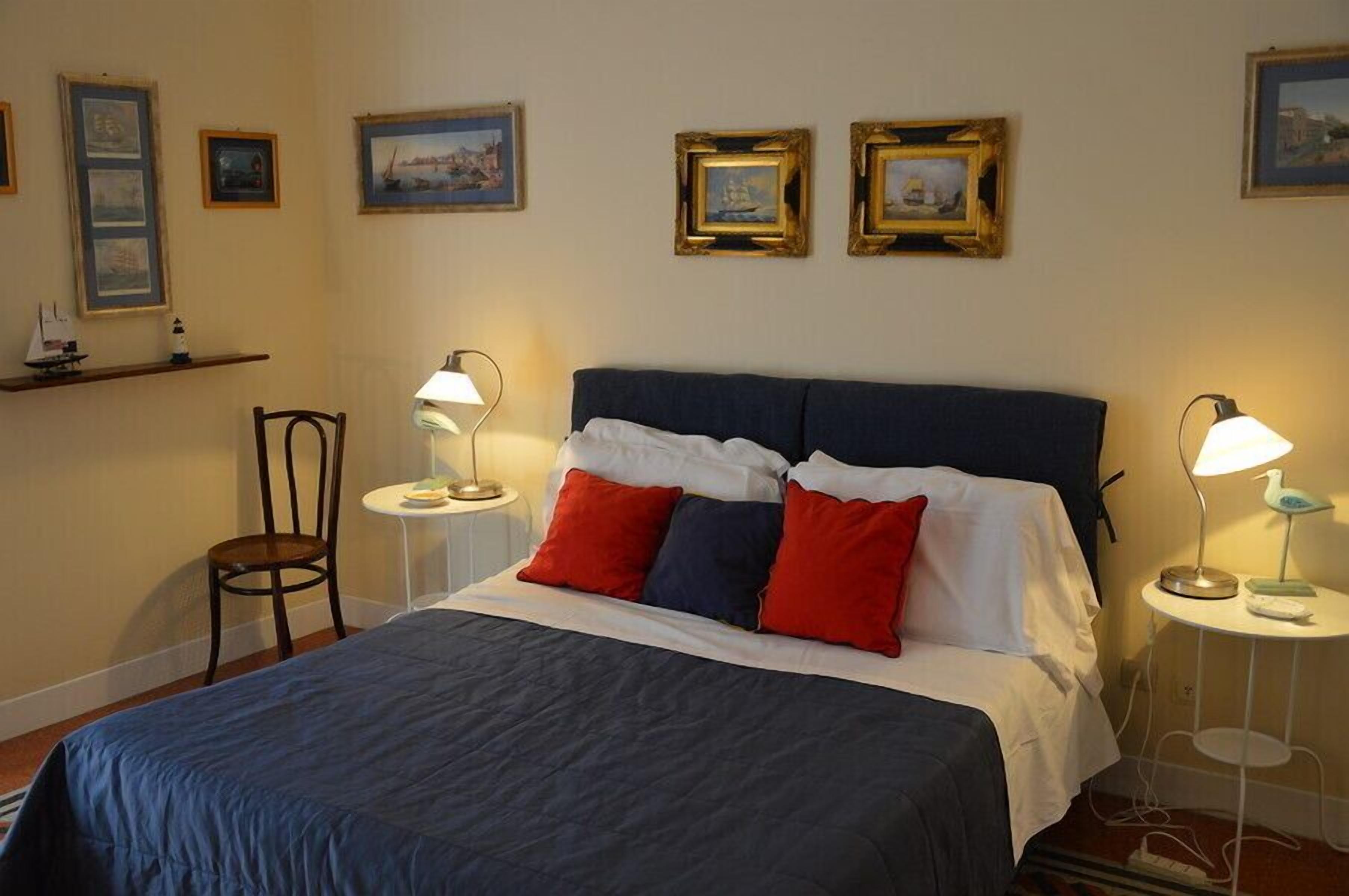 A neatly made bed with a blue and white bedspread, two red pillows, and a dark blue headboard. There are two bedside tables with lamps, a chair, and framed pictures on the wall.
