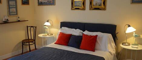 A neatly made bed with a blue and white bedspread, two red pillows, and a dark blue headboard. There are two bedside tables with lamps, a chair, and framed pictures on the wall.