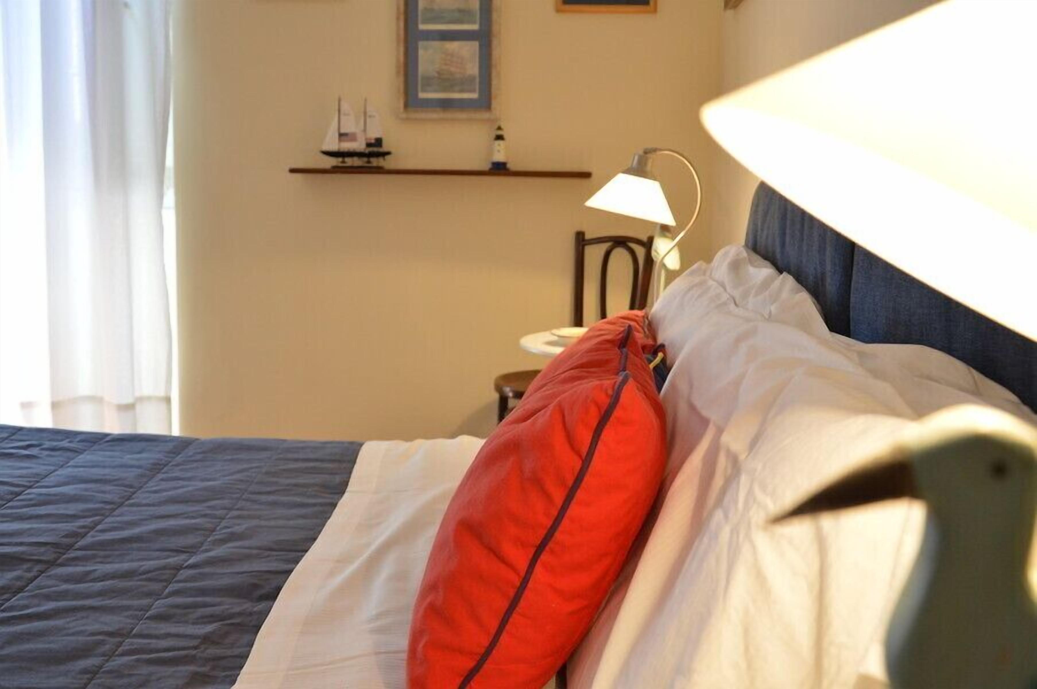 Un lit bien fait, agrémenté d’un oreiller rouge, d’un oreiller blanc et d’une tête de lit bleue. On aperçoit, à l’arrière-plan, une petite table avec une lampe et une étagère où sont disposés des objets décoratifs.