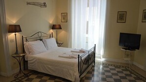 Una cama bien hecha con ropa de cama y toallas blancas, un suelo a cuadros, un televisor sobre un mueble bajo y dos lámparas sobre mesitas de noche.