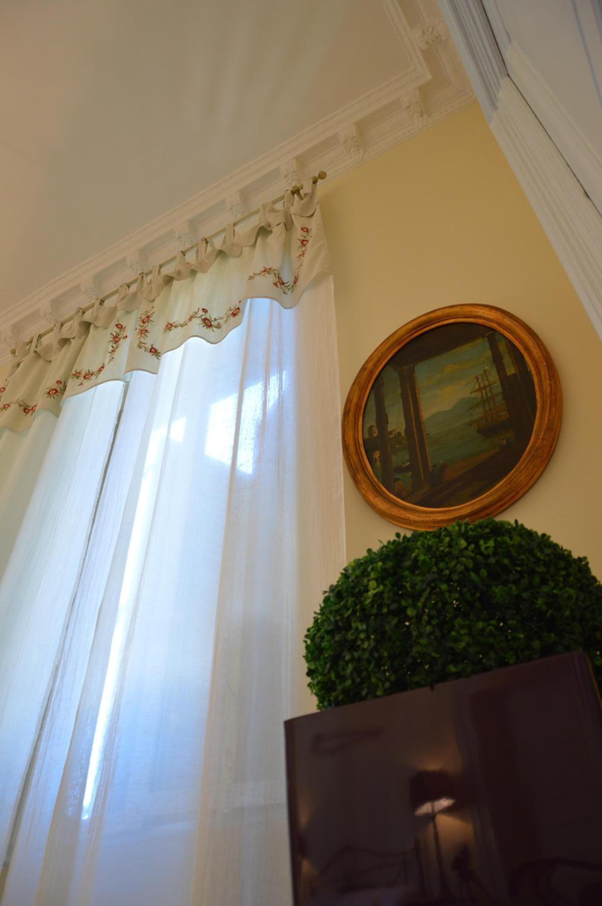 Une pièce dotée d’une grande fenêtre ornée de rideaux légers et d’une lambrequin à motifs floraux, d’un miroir rond accroché au mur et d’une plante en pot placée sous le miroir.