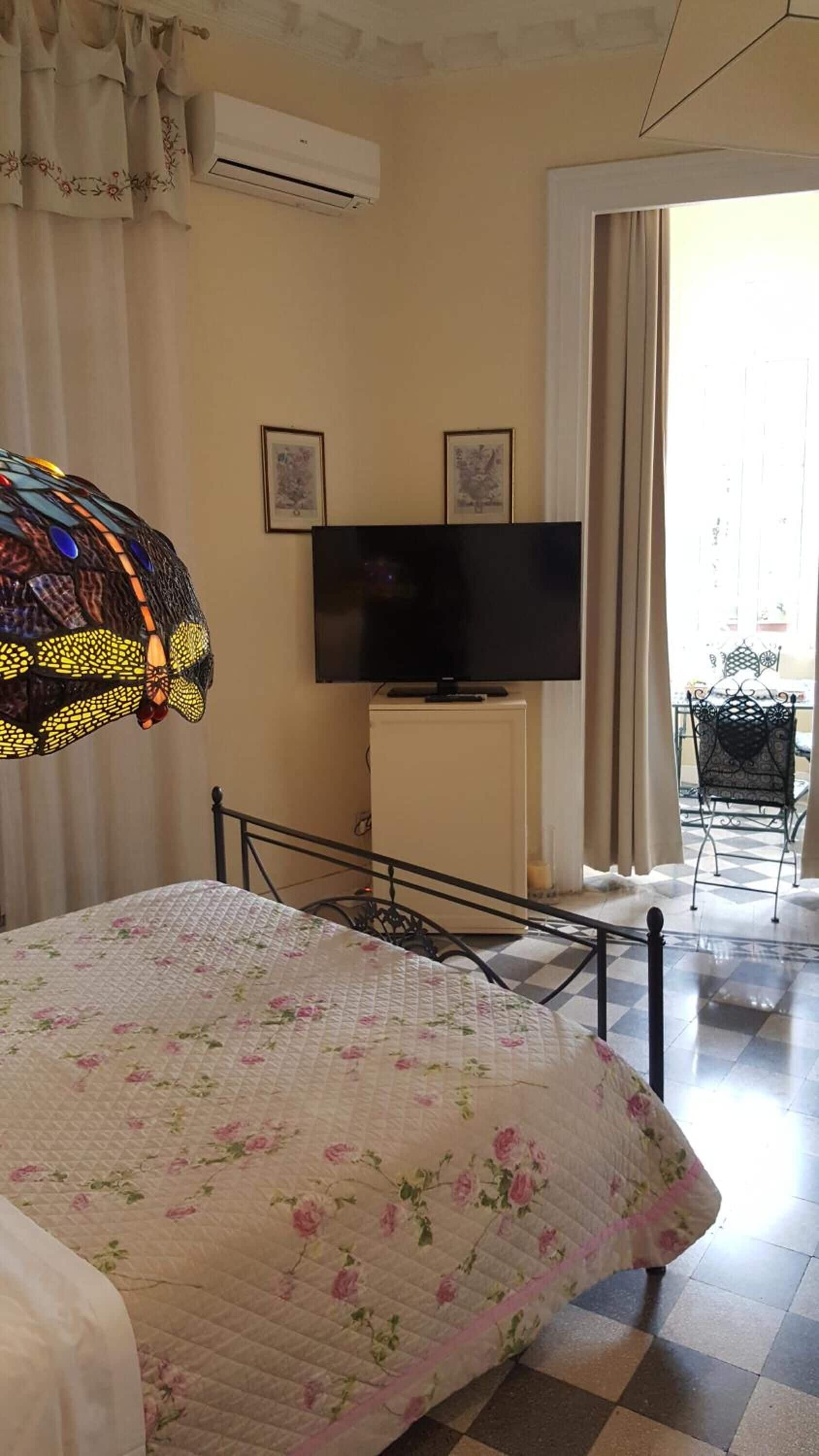 A hotel room with a bed, a television on a cabinet, a window with curtains, and a balcony with a chair.