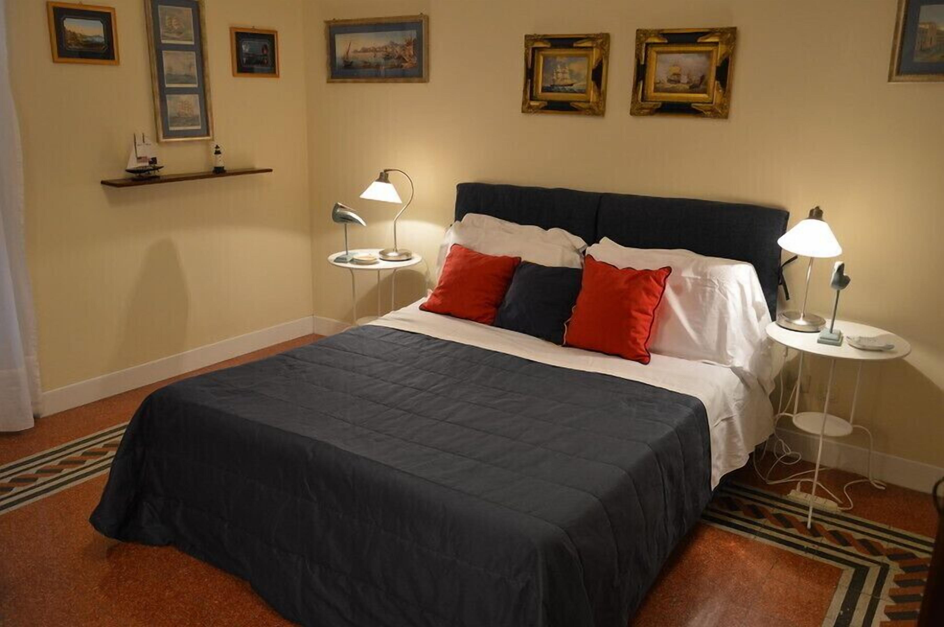 Een netjes opgemaakt bed met een donkere sprei en rode en zwarte kussens, twee nachtkastjes met lampen, en een klein plankje met decoratieve objecten.