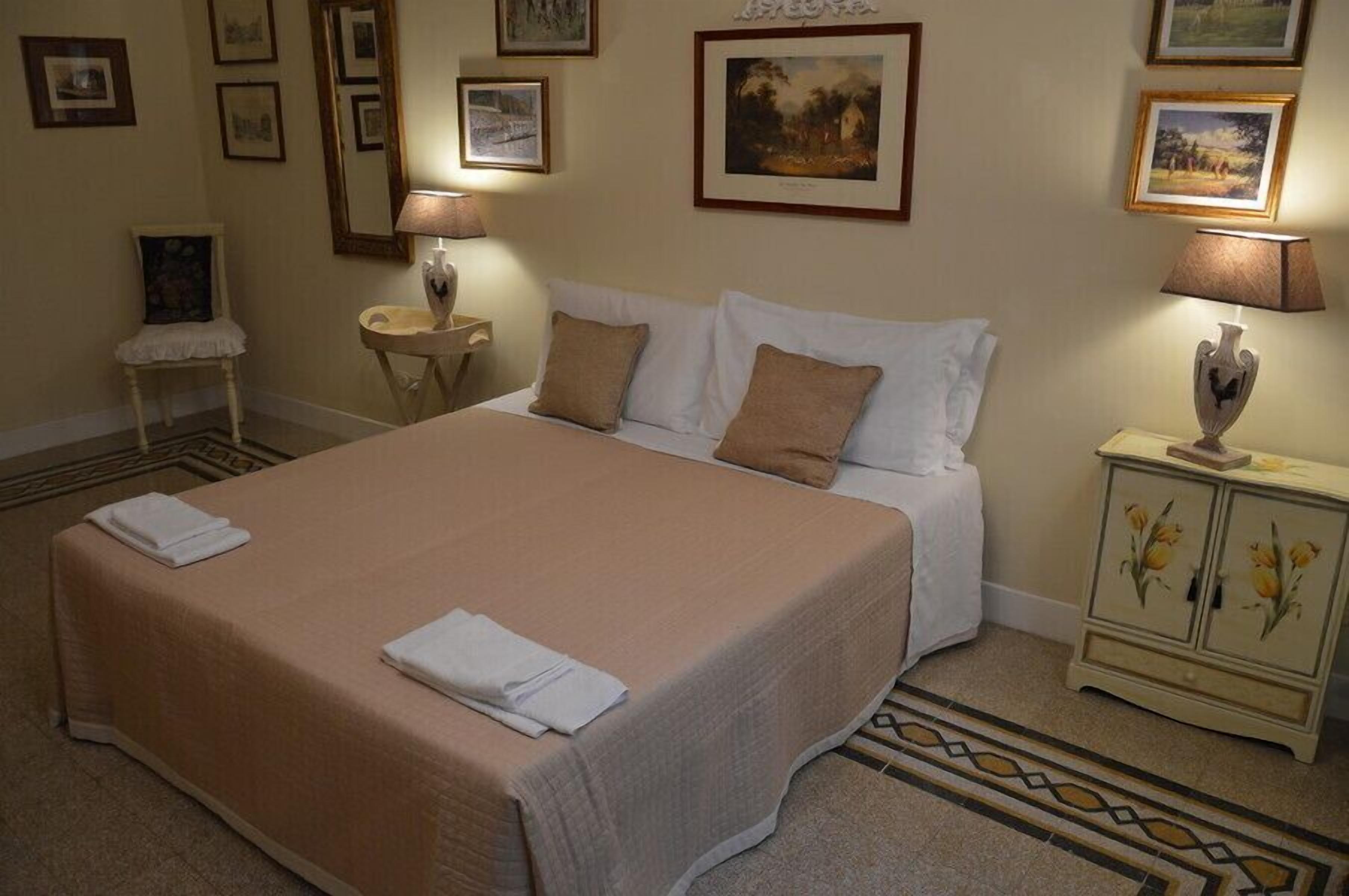 A neatly made bed with a beige bedspread and white pillows, a small bedside table with a lamp, and a cabinet with floral designs.