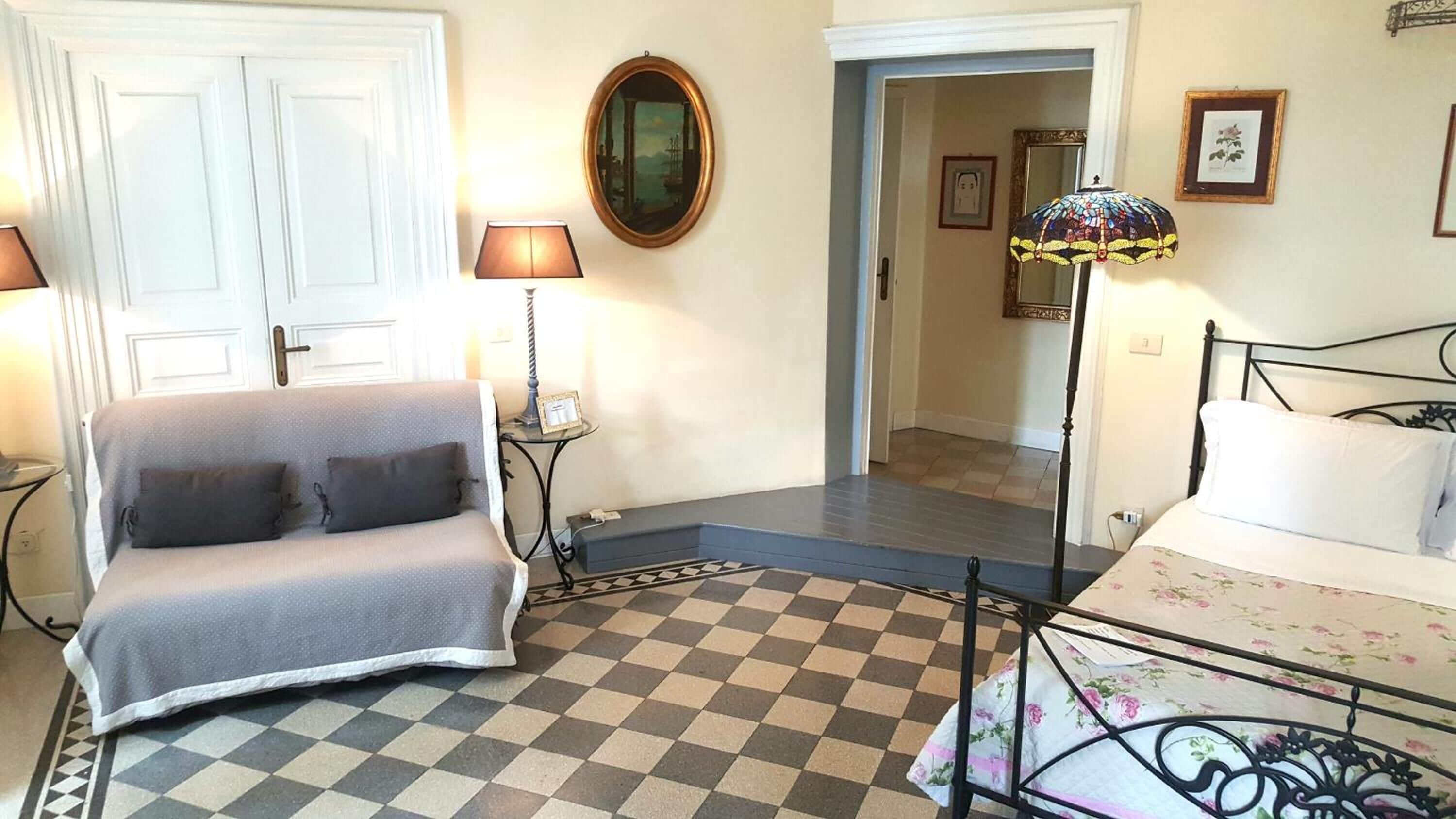 A room with a checkered floor, a grey sofa, a bed with floral bedding, a mirror, and a lamp.