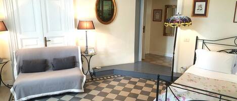A room with a checkered floor, a grey sofa, a bed with floral bedding, a mirror, and a lamp.