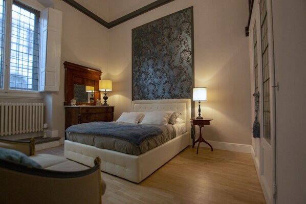 A bedroom with a large bed, a nightstand, a dresser, a window with blinds, and a large abstract painting on the wall.