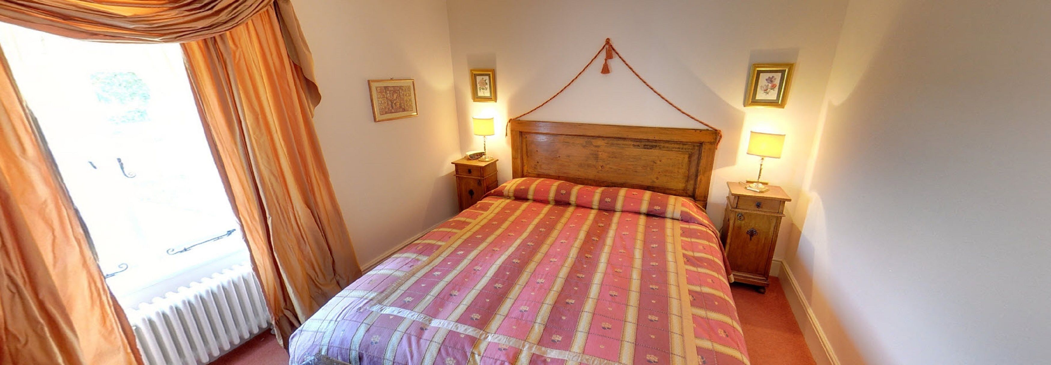 A bedroom with a wooden headboard, a bed with a patterned bedspread, two bedside tables with lamps, and a window with curtains.