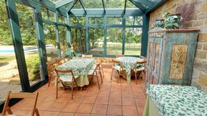 A conservatory with a glass roof, wooden chairs, and tables covered with patterned tablecloths. - Brampton Grange (Northampton)