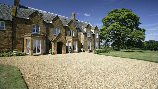 A stone building with a gravel driveway and a large tree in the background. - Brampton Grange (Northampton)