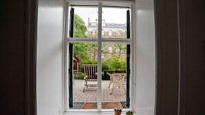 Vista por una ventana con marco blanco, mostrando un edificio de ladrillo, una silla de madera y una pequeña mesa con plantas en macetas.