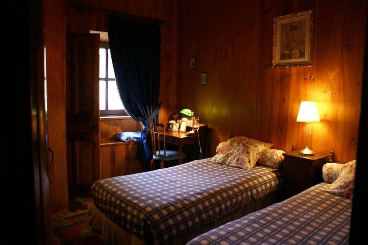 A cozy room with two beds, a desk with a lamp, a window with curtains, and a framed picture on the wall.