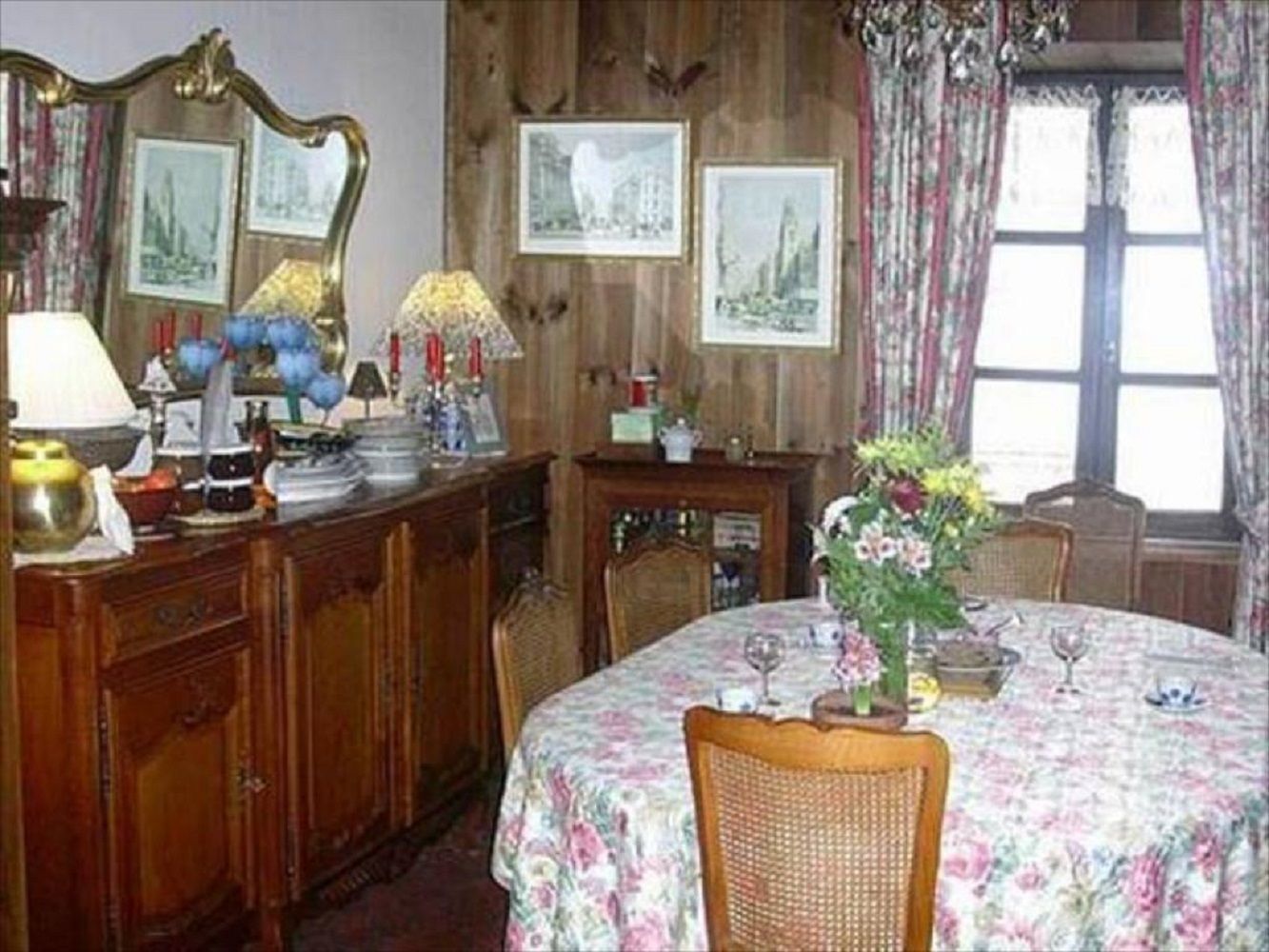A dining area with a round table, chairs, a floral tablecloth, a vase with flowers, and framed pictures on the wall.