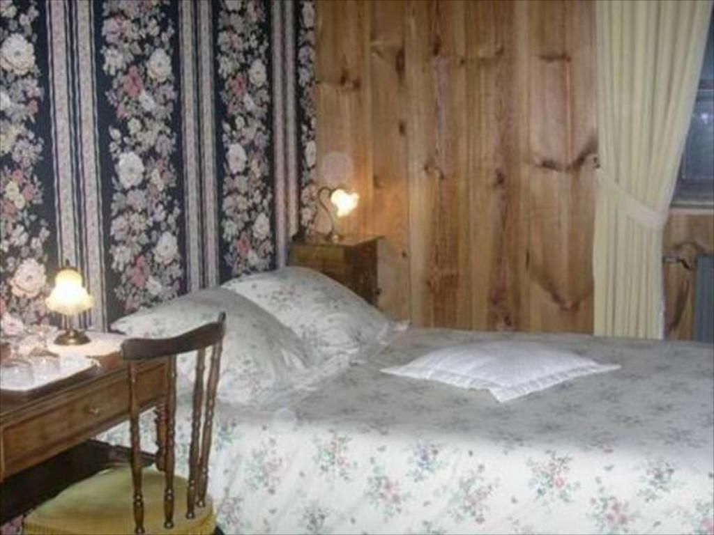 A bedroom with a wooden wall, a floral bedspread, a bedside table with a lamp, and a chair.