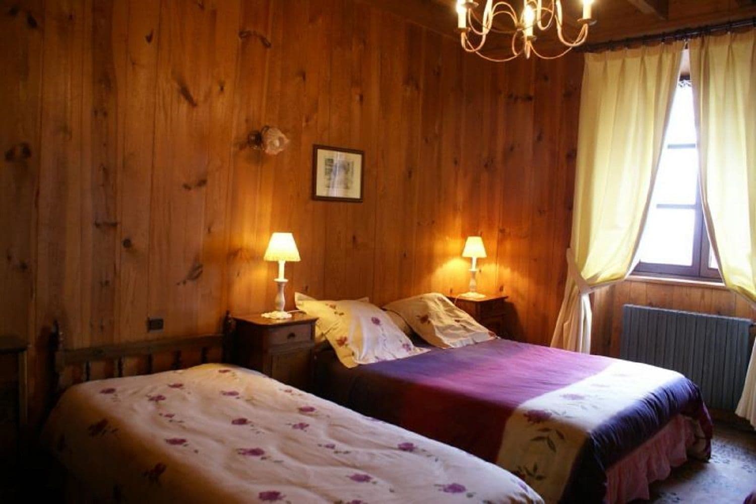A wooden room with two beds, each with a bedside table and lamp, a window with curtains, and a chandelier.