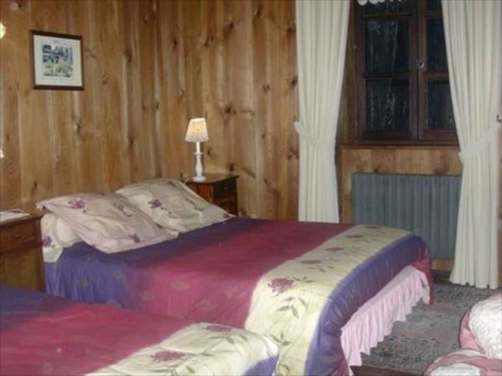 Une chambre avec des murs en bois, un lit recouvert d’une literie à motifs floraux, une table de chevet avec une lampe et une fenêtre avec des rideaux.