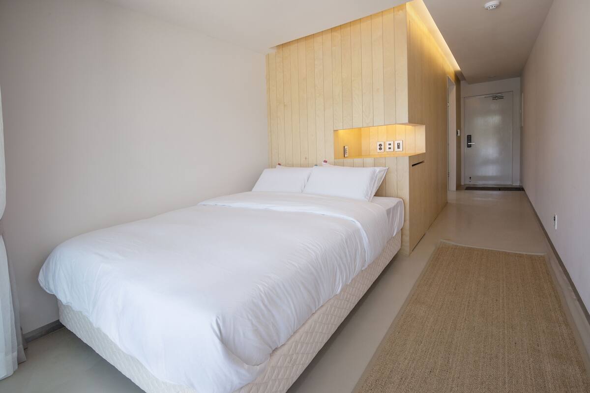 A hotel room with a large bed, wooden headboard, bedside lamp, and a door leading to a hallway.
