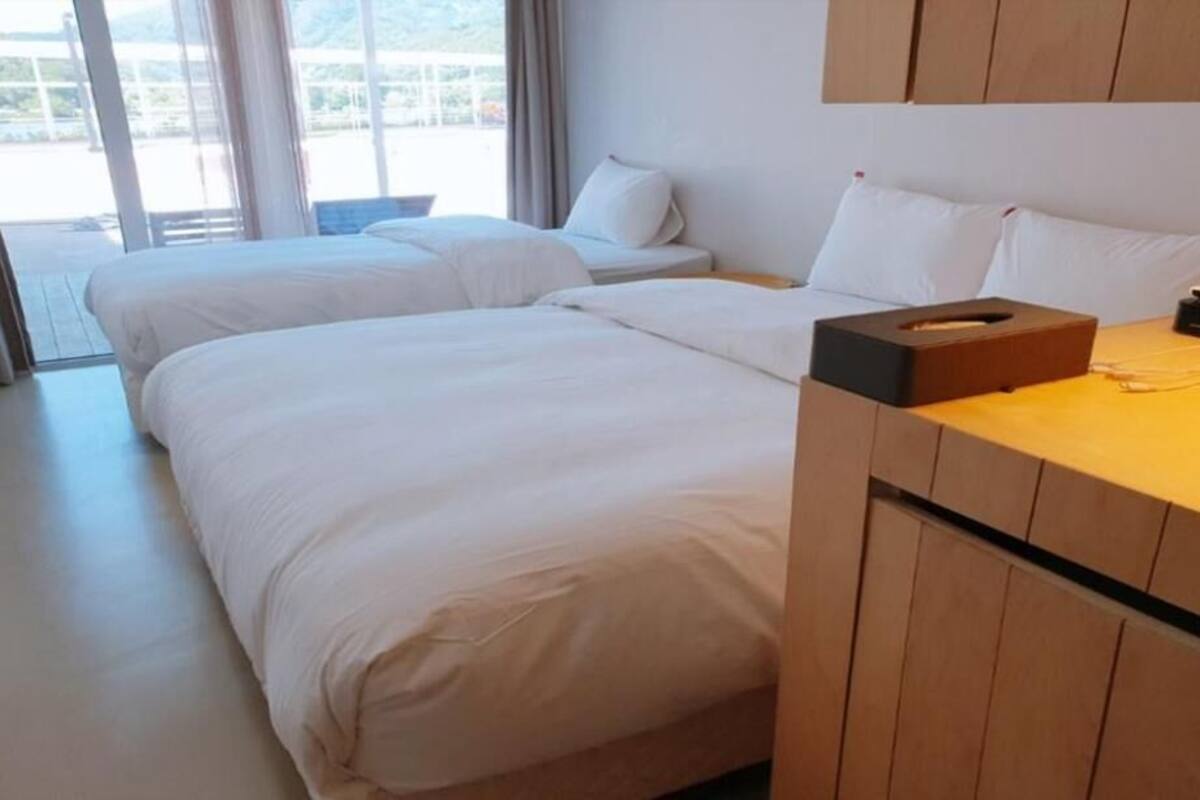 A hotel room with two beds, a wooden headboard, a bedside table with a tissue box, and a view of the outdoors.