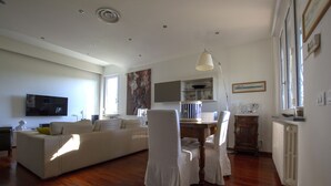 Uma sala de estar ampla com um sofá branco grande, uma mesa de jantar com cadeiras e uma área de cozinha ao fundo.