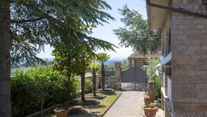 Uma passagem leva a um edifício de pedra com telhado de telhas, cercado por vasos de plantas e árvores.