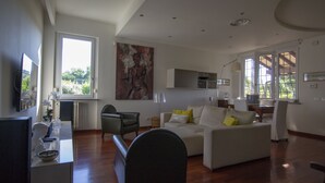 Uma sala de estar ampla, com sofá, cadeiras e uma mesa de jantar. Há uma janela grande que traz luz natural e vista para o verde l á fora.