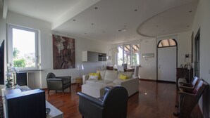 Uma sala de estar espaçosa com um sofá grande, uma mesa de centro e uma televisão. Há uma porta que dá para outro cômodo e uma janela que proporciona iluminação natural.