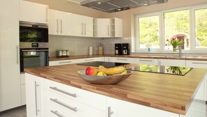 A modern kitchen with white cabinets, a stainless steel range hood, a built-in oven, a large island with a bowl of fruit, and a window with a view of greenery. - Villa Thormarcon (Malchow)