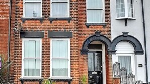 Una casa de ladrillo de dos pisos con entrada principal, ventanas grandes y un pequeño jardín con plantas en macetas.