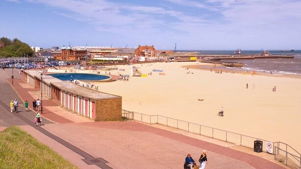 Una playa con piscina, paseo marítimo y edificios al fondo.