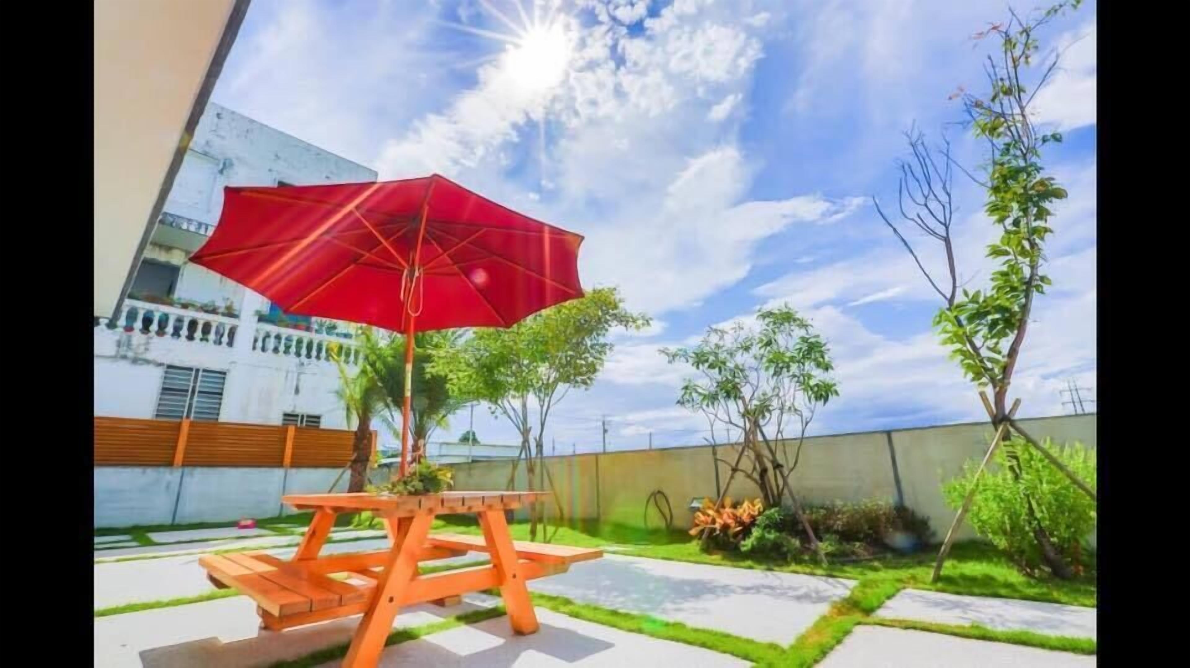 An outdoor patio with a wooden table and a red umbrella, surrounded by greenery and a white building in the background.