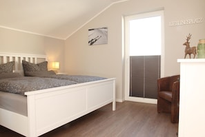 A bedroom with a white bed, a nightstand with a lamp, a window with blinds, and a wall-mounted shelf with decorative items. - Bergtraum Braunlage (Braunlage)