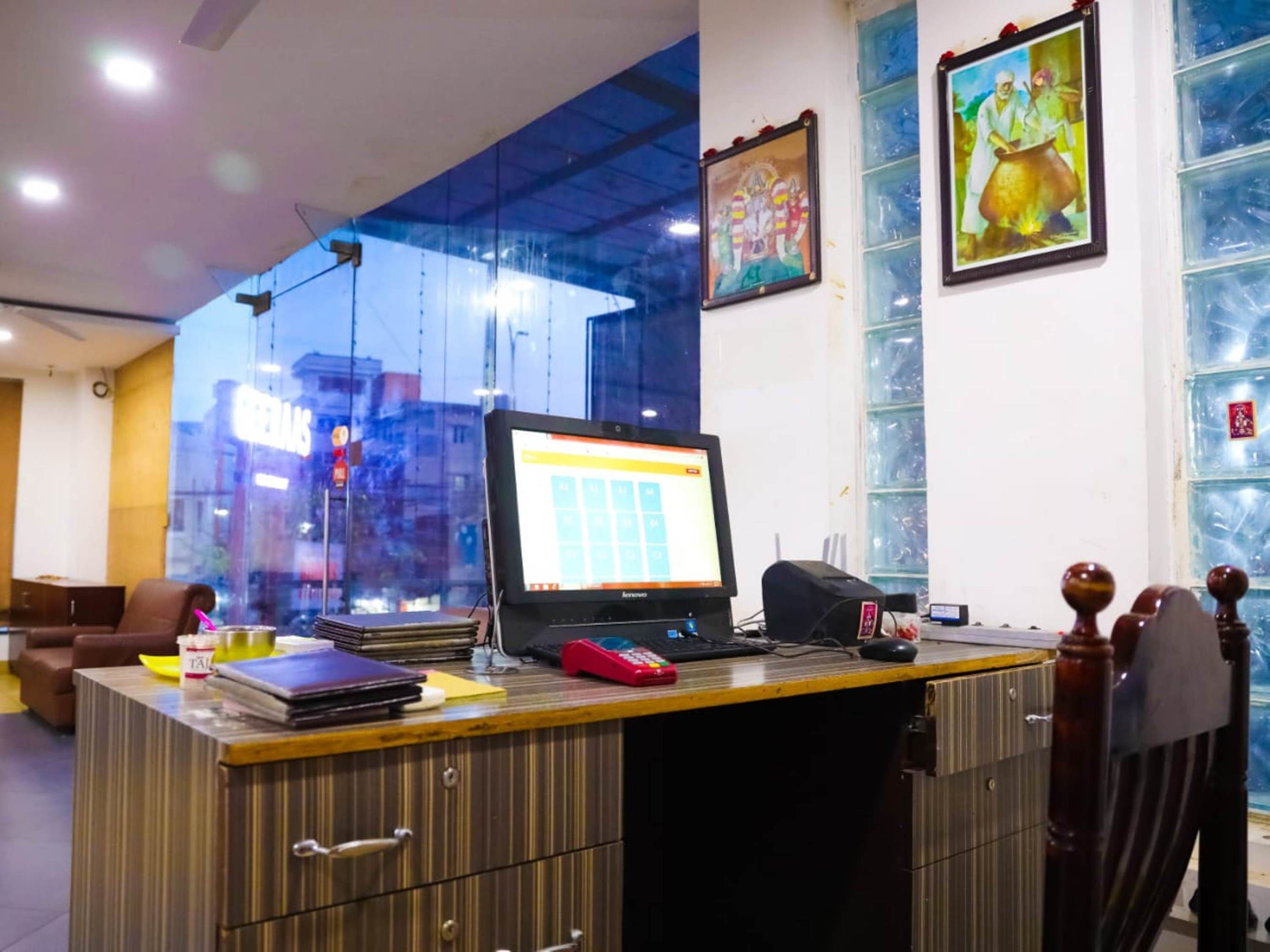 A reception desk with a computer monitor, a telephone, and a cash register. There are framed pictures on the wall and a window with a view of the city.
